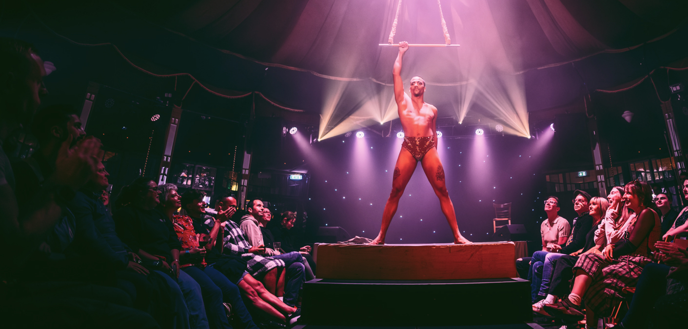 An aerialist holding a trapeze centre stage in a spiegeltent with an audience looking on