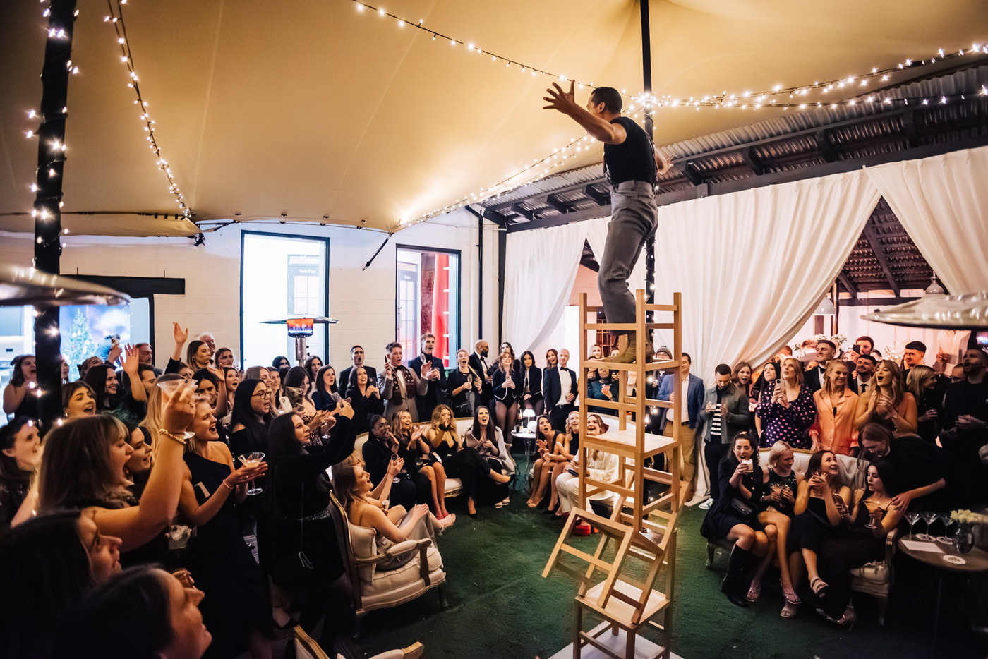 A variety performer standing atop a tall stack of chairs with an audience looking on at a corporate event