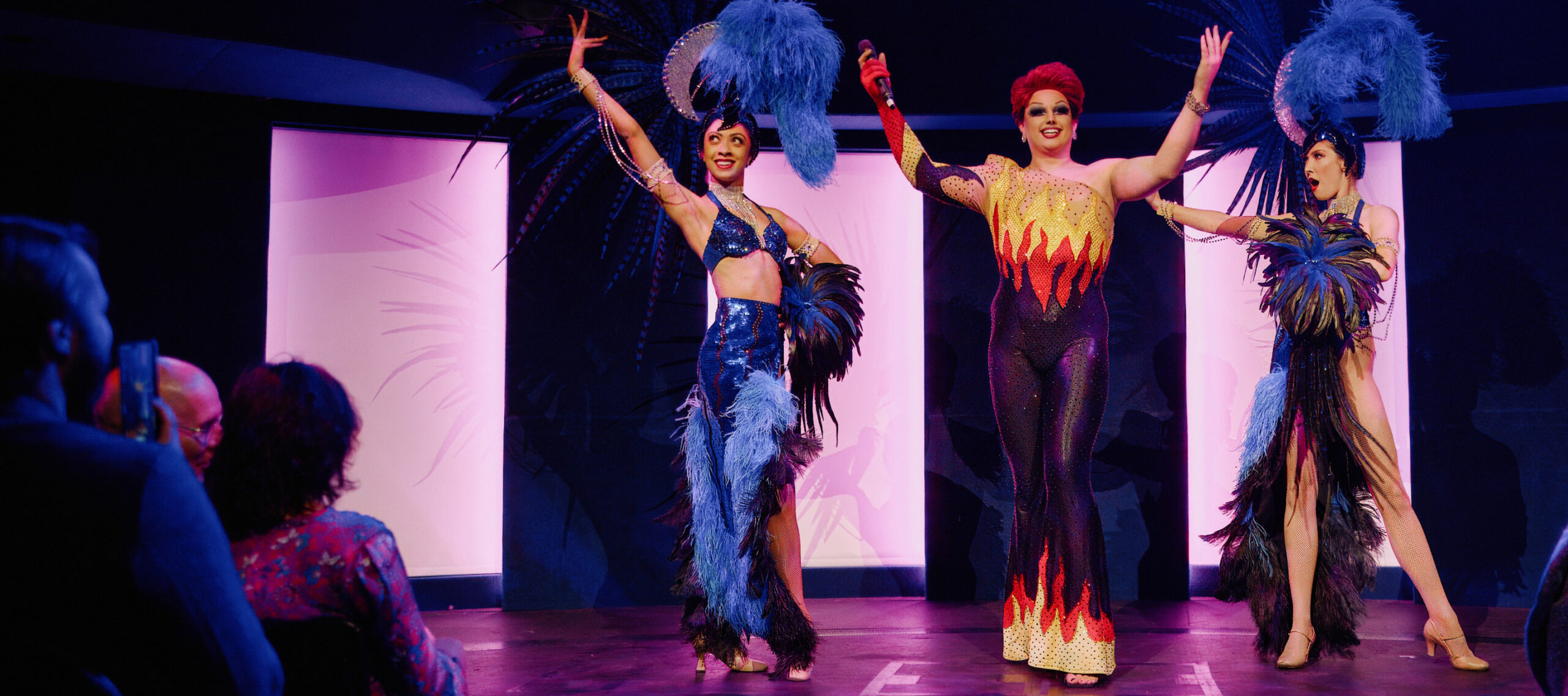 A drag artist supported by two showgirls opening a variety and cabaret show at Underbelly Boulevard